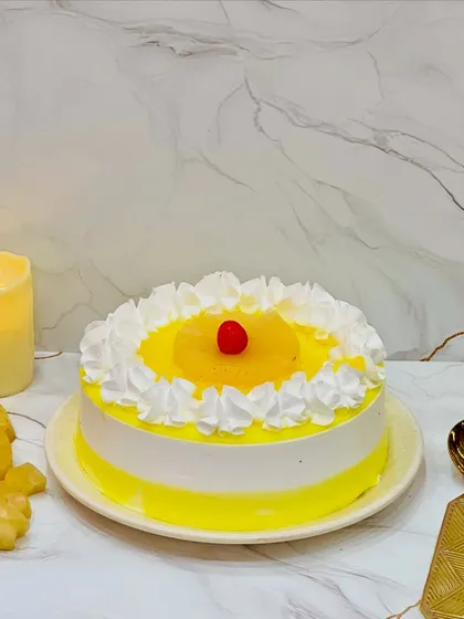 A classic Pineapple Cake, beautifully presented. This cake features a soft vanilla sponge, pineapple chunks, and a light cream frosting, topped with a pineapple slice and a cherry. It's a timeless favorite for any family gathering.