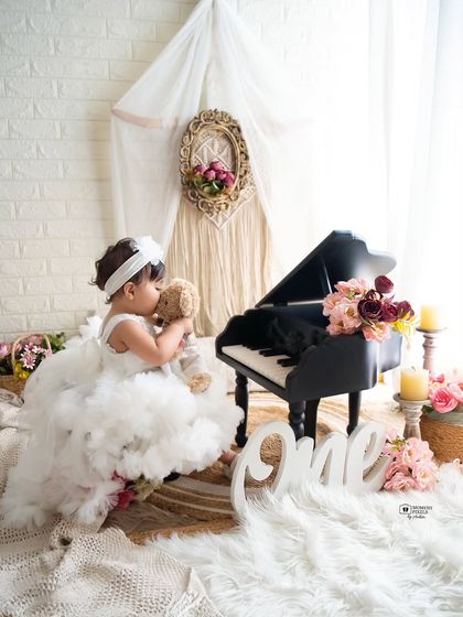 A dreamy first birthday scene with a little pianist. Dressed in a fluffy white gown, she shares a sweet moment with her teddy bear, creating a timeless memory of turning one.