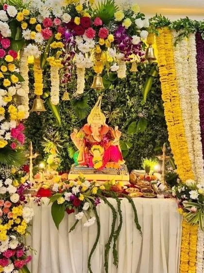 This rich and colorful Ganpati decor combines multiple flower types and colors, including marigolds, orchids, and roses. The dense, multi-layered arrangement with hanging bells creates a truly grand and festive atmosphere.