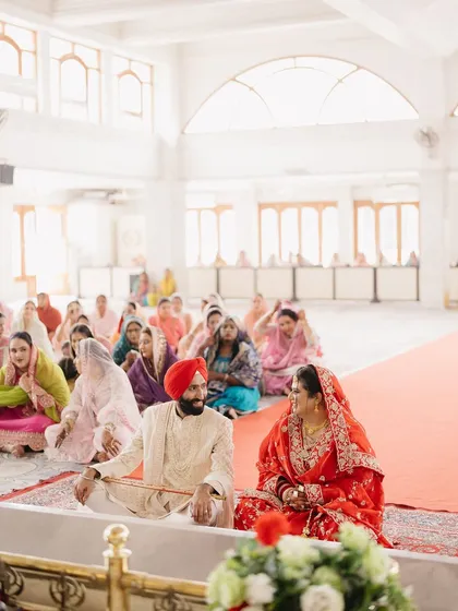 A wide shot of the Anand Karaj ceremony, showing the couple seated before the Guru Granth Sahib. The image captures the peaceful and communal nature of the Sikh wedding.