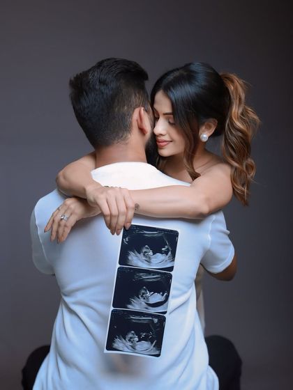 A playful and intimate announcement shot. The mom-to-be hugs her partner from behind, showcasing a strip of sonogram photos on his back, symbolizing their journey together.