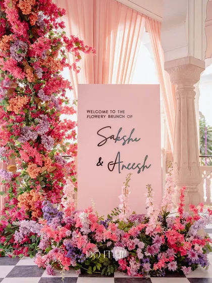 A beautiful welcome sign for a "Flowery Brunch" in Jaipur. The soft pink sign is framed by a stunning, overflowing arch of bougainvillea and other flowers in shades of pink, orange, and red.