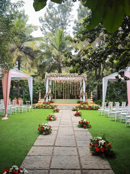 A classic wedding ceremony setup with a floral mandap and flower petals lining the aisle. The tall palm trees in the background add to the tropical, garden feel.