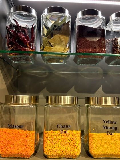 A close-up showing whole spices like red chillies and bay leaves stored alongside various dals. The clear jars make it easy to distinguish between masoor, chana, and moong dal.