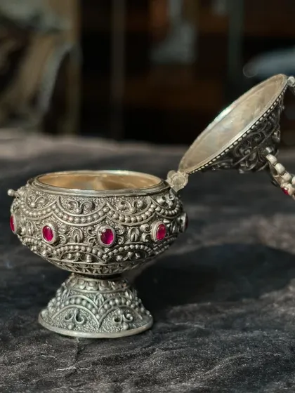 The silver kumkum box is shown open, revealing the container inside. The intricate carving on the base and the fine details of the hinged lid showcase its excellent craftsmanship.