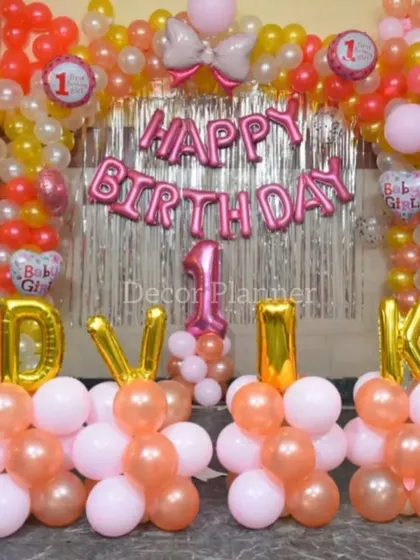 A lovely first birthday decoration at home for Advika. The setup features a balloon arch in pink and peach, a shimmer curtain backdrop, and her name spelled out in letter balloons.