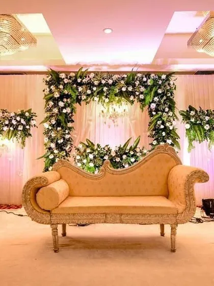 For a more classic and sophisticated look, I designed this engagement stage with a simple beige divan couch. The backdrop features an arch of white flowers and greenery against soft drapery, illuminated by elegant chandeliers.