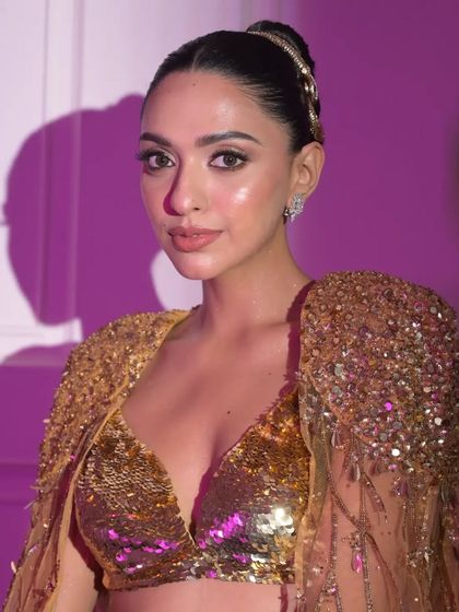 A portrait showing the front view of the look. The hair is styled in a severe, high bun, which complements the bold makeup and sequined gold outfit for a powerful editorial statement.