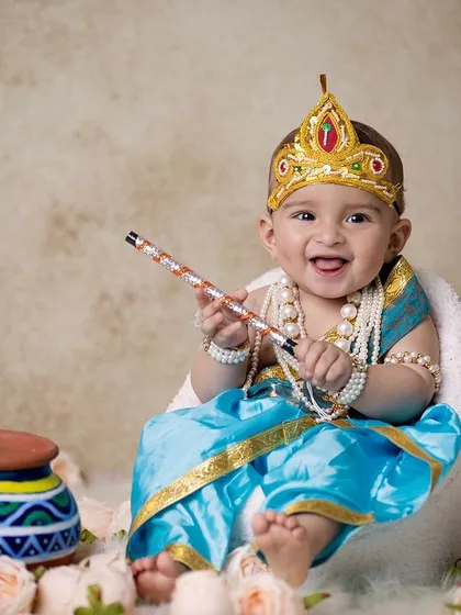A joyful baby Krishna holding his flute, seated in a mini armchair surrounded by flowers. The lighting and setup create a warm and inviting portrait.