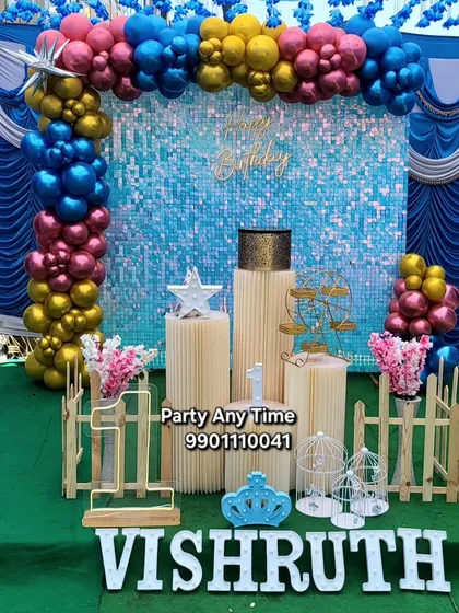 A vibrant first birthday setup for Vishruth with a blue shimmer wall and a colorful balloon arch. The light-up name and number add a special touch.