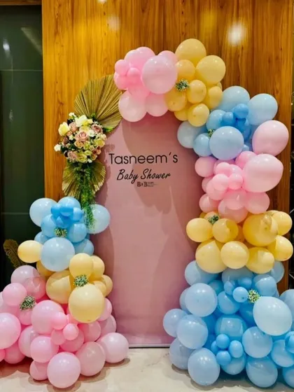 A personalized baby shower setup with a pastel balloon arch in pink, blue, and yellow. The custom pink backdrop features the mother's name, and the arrangement is accented with dried palm leaves and flowers.