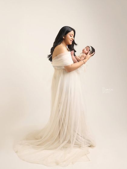 A full-length portrait showcasing the grace of new motherhood. The mother, dressed in a flowing white gown, cradles her newborn, creating a timeless and artistic image of their connection.