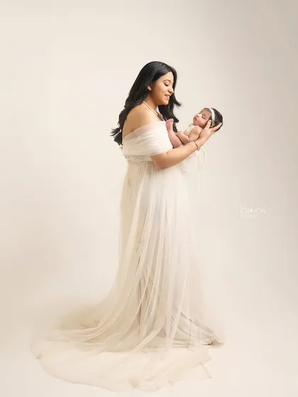 A full-length portrait of a mother in a flowing white gown, holding her newborn. It showcases the elegance and grace of new motherhood.