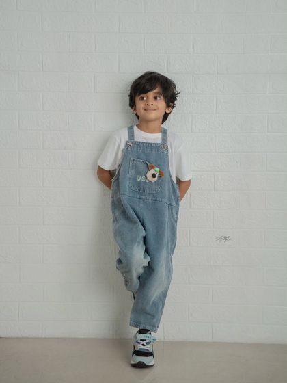 A simple and classic portrait of a young boy in denim overalls leaning against a white brick wall.