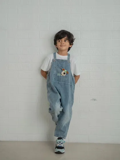 A simple and classic portrait of a young boy in denim overalls leaning against a white brick wall.