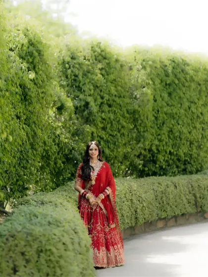 A full-length bridal portrait in a garden setting, the bride's red lehenga standing out beautifully against the green foliage.