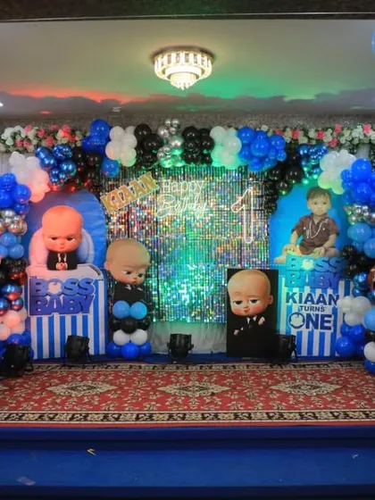 A full stage setup for a Boss Baby first birthday in a banquet hall, complete with a sequin backdrop and multiple character props.