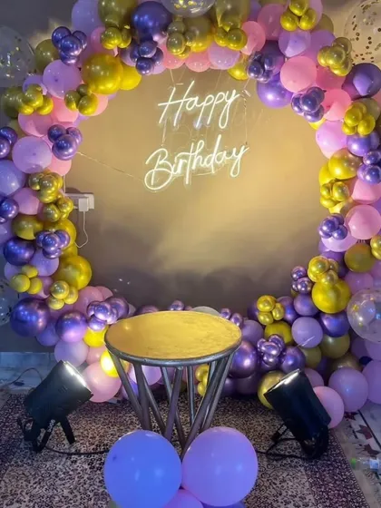 A circular balloon photo booth in shades of purple, pink, and gold. The "Happy Birthday" neon sign in the center makes it the perfect spot for party pictures.