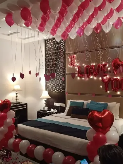 A romantic decoration with 'I Love You' foil balloons and a bed framed by pink and white balloon pillars. The ceiling is covered with balloons with hanging ribbons.