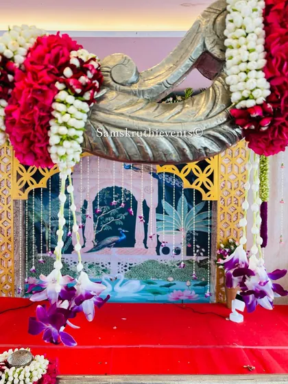 A close-up of a fusion backdrop, where a traditional peacock painting is framed by a silver carved arch and hanging floral garlands.