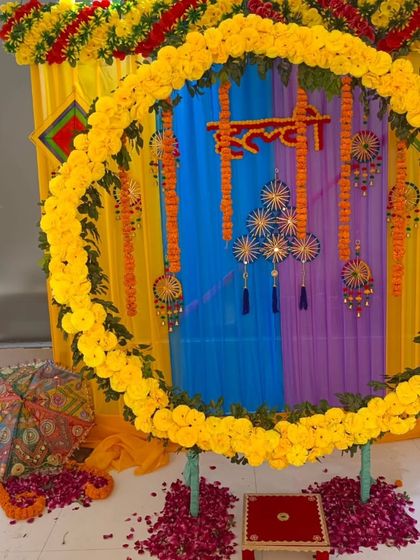 A colorful Haldi setup with a circular marigold backdrop. The decor includes a small stool for the ceremony, Rajasthani umbrellas, and a floor scattered with rose petals.