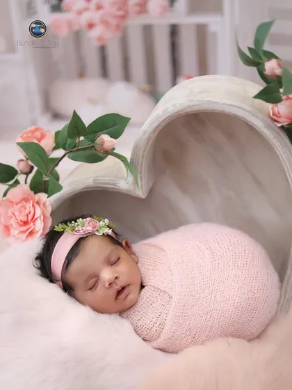 A newborn girl, swaddled in pink, sleeps inside a heart-shaped wooden bowl surrounded by delicate pink roses, creating a truly romantic and sweet portrait.