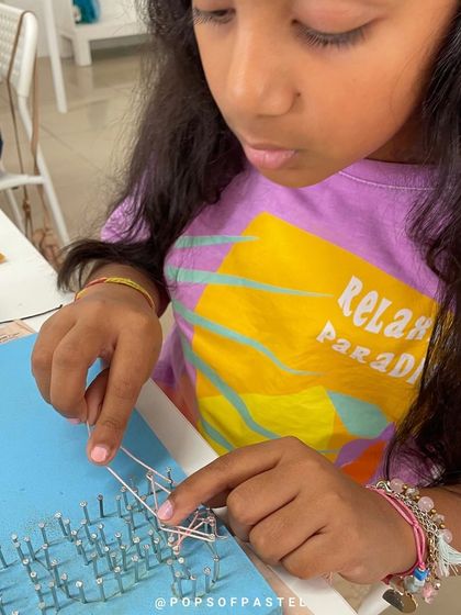 A young girl learns the intricate process of weaving string between nails, developing fine motor skills and patience.