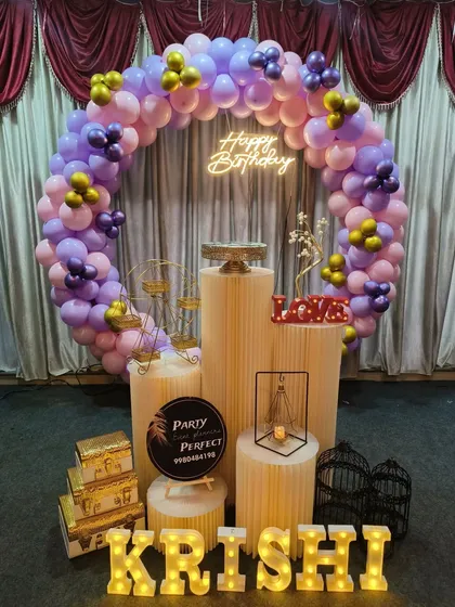 A pretty pastel balloon ring in shades of purple and pink for Krishi's birthday. The setup includes a neon sign, light-up name, and various decorative props on pedestals.
