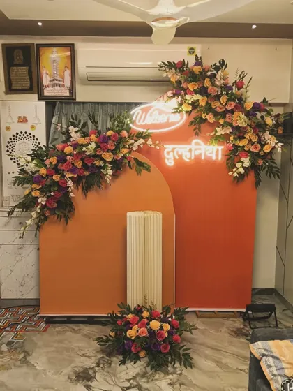 A vibrant "Welcome Dulhaniya" (Welcome Bride) setup for a home celebration. The bright orange arched backdrop is decorated with colorful flowers and a white neon sign.