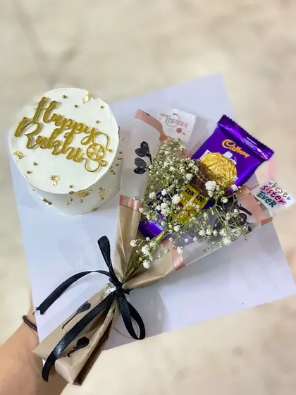 An elegant Rakhi gift set featuring a white bento cake with "Happy Rakhi" written in beautiful gold script, paired with a classic chocolate and baby's breath bouquet.