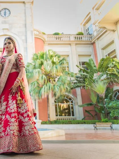 A breathtaking full-length shot of a bride in her red lehenga, standing in the courtyard of a European-style luxury hotel.