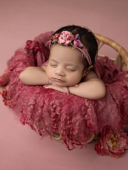 A baby girl rests her chin on her hands in a basket filled with soft, textured pink fabric. The floral headband adds the perfect finishing touch.