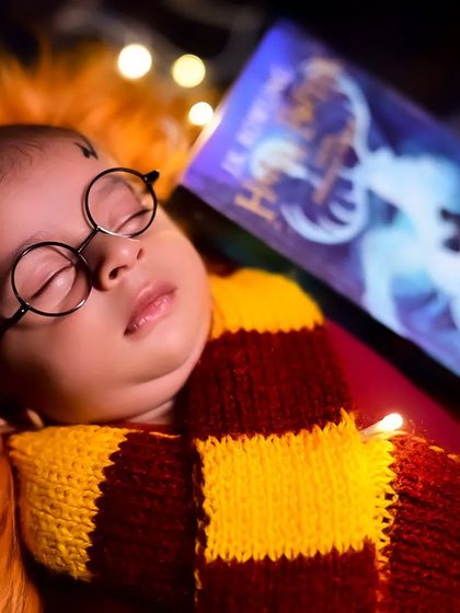 A close-up of a newborn dressed as Harry Potter, sleeping soundly with a book. The soft lighting and props create a magical scene.