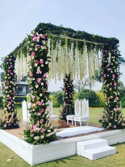 An elegant outdoor wedding mandap for a daytime ceremony, featuring pillars wrapped in lush pink and white florals, with a ceiling of hanging white wisterias creating a romantic and natural setting.