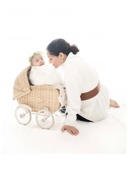A mother leans over to gaze lovingly at her newborn, who is resting in a charming wicker pram. This clean, high-key studio shot is full of tenderness and captures the quiet admiration of a mother for her new baby.