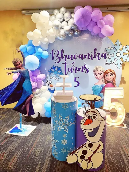 A 'Frozen' themed birthday party featuring Elsa, Anna, and Olaf. The decor includes a character-filled backdrop, snowflake details, a light-up number '5', and a balloon arch in icy shades of blue, purple, and silver.