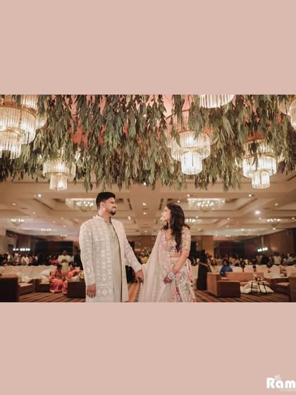 A wide shot of the couple holding hands under a ceiling adorned with lush greenery and chandeliers, capturing the grand decor of their engagement venue.