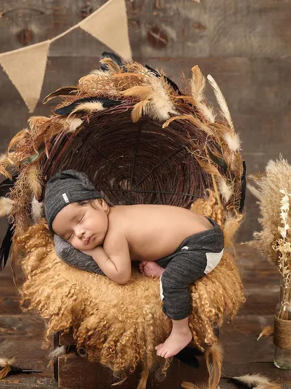 A newborn sleeps on a fluffy surface in a nest-like basket decorated with pampas grass and feathers, perfectly capturing a boho-rustic aesthetic.
