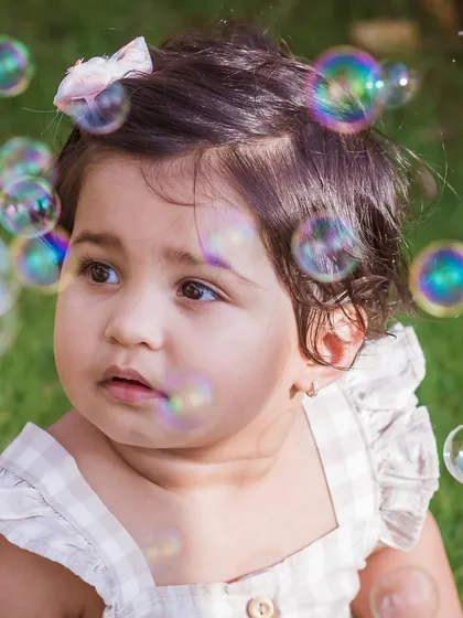 A close-up portrait of a baby girl looking thoughtfully at floating bubbles. The soft focus and dreamy quality of the image make it feel magical.