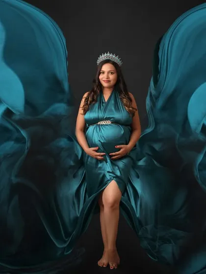 A portrait that commands attention. The mom-to-be stands as a queen in a teal gown with fabric flowing like wings, accented with a delicate crown.