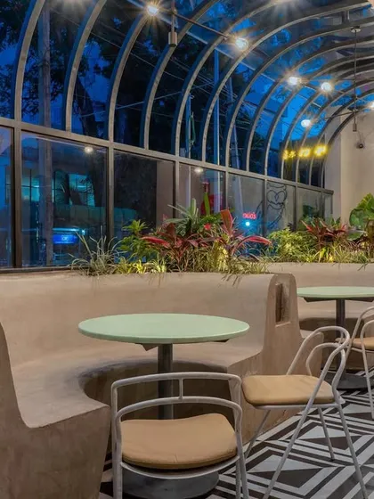 The semi-outdoor seating at Hangover is housed under a vaulted glass ceiling, creating a greenhouse-like effect. The space combines built-in concrete seating with lush planters and bold geometric floor tiles.