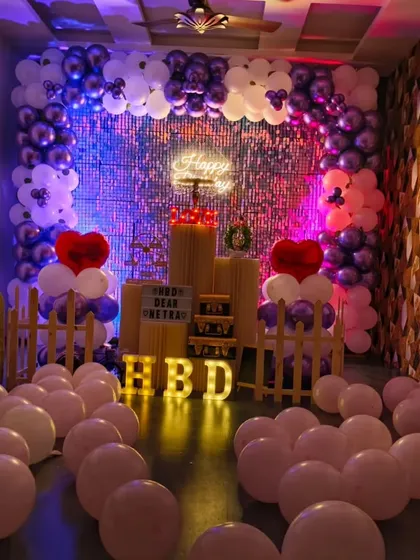 The purple and white birthday setup seen with different colored lighting. The shimmering backdrop reflects the lights beautifully, creating a dynamic and festive atmosphere.