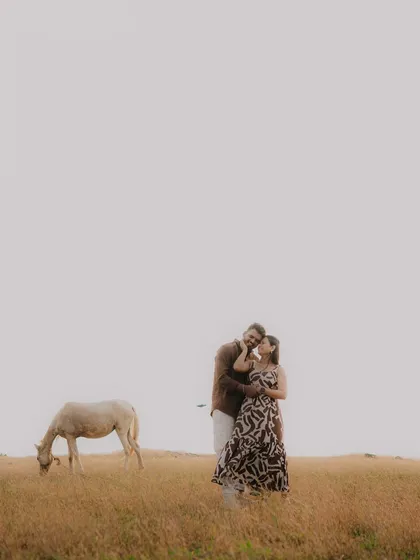A wide, minimalist shot of a couple in a vast, golden field, with a horse grazing nearby. This image has a peaceful, storybook quality, emphasizing the couple's connection within a beautiful, expansive landscape.
