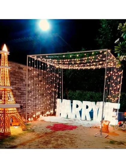 This is my signature grand proposal setup, arranged in a garden at night for maximum impact. It features large, illuminated 'MARRY ME' letters under a canopy of draped fairy lights, with rose petals on the ground to create a truly romantic pathway.