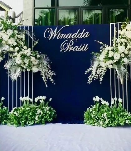 A close-up of a chic engagement stage. The deep blue backdrop with custom white calligraphy, framed by modern white structures and floral arrangements, creates a bold and personalized statement.