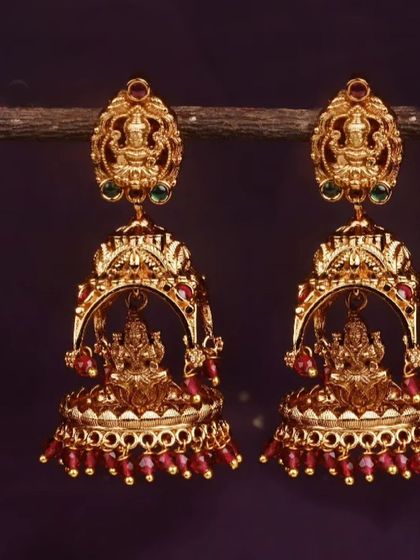 This pair of matte gold finish jhumkis features a Goddess Lakshmi design and is finished with rich ruby-red bead drops. Perfect for adding a pop of color to your festive attire.