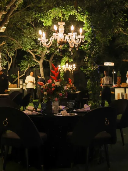 An outdoor lounge area for a monochrome-themed party, where chandeliers hang from trees over elegant black dining tables.