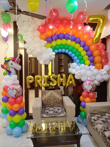 A detailed rainbow and unicorn party setup in a living room. The design features a large rainbow arch built around a doorway, with balloon columns, a unicorn figure, and the name "PRISHA" in gold foil letters.