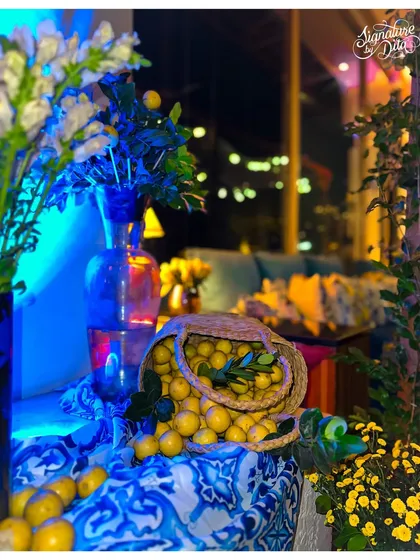 This decor detail shows a woven basket overflowing with fresh lemons, set against a blue patterned cloth and illuminated by cool blue light, capturing the essence of Mediterranean charm.