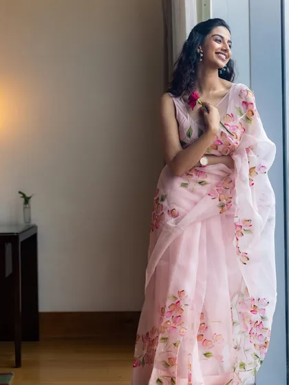 A full-length shot of the pink organza saree by a window. The natural light beautifully illuminates the sheer fabric and delicate floral painting, creating a soft and ethereal mood.
